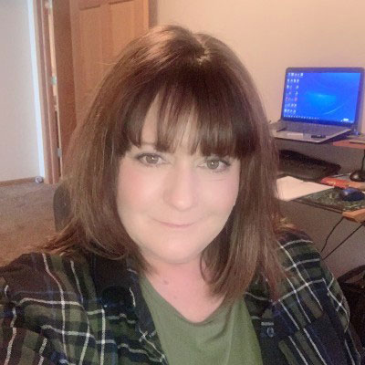 Katrina Patton has shoulder-length brown hair and bangs, wearing a green shirt and plaid flannel jacket, taken in a home office setting. 