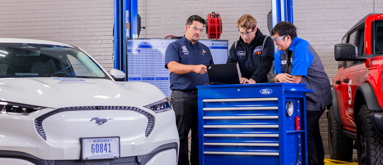 Mark Berggren and two Ford ASSET students are reviewing diagnostic information on a laptop on top of a blue tool cart. Two are wearing dark uniforms with safety glasses, and one is wearing a blue shirt. A white Mustang and red Bronco surround them. 