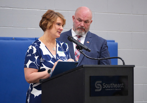 Dr. Bobbi Berner speaks at the Scholarship Luncheon in a blue and white dress with Jason Berner in a suit looking on. 