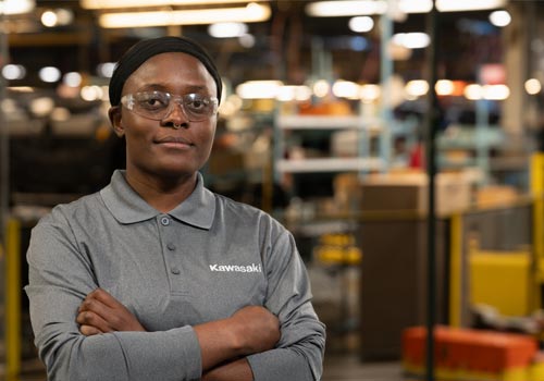 Benie Buzi Mbo Nta stands in a maintenance shop in a grey polo with the Kawasaki word mark on the left chest.
