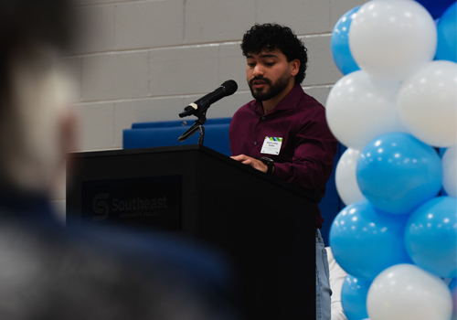 Roberto Paredes speaks at the SCC Educational Foundation Scholarship Luncheon. He is in a maroon shirt  with blue and white balloons visible on the left side.