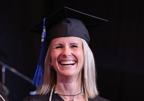 Crissy Murphy smiles at the 2025 Spring Graduation ceremony wearing a black graduation cap with a blue tassel against a dark background.