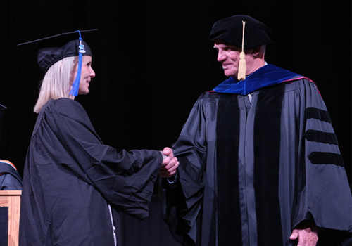 Crissy Murphy (left) in a black cap and gown shakes hands with SCC President Dr. Paul Illich wearing formal academic attire with blue trim during the May 14, 2025 graduation ceremony on a darkened stage.