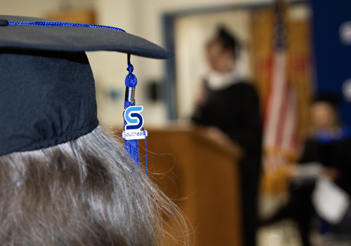 A close-up view of a mortarboard with a blue tassel featuring the Southeast Community College logo medallion, worn by a graduate with gray hair. The photo is taken from behind as Dean Amy Doty is blurred at the podium and an American flag visible in the background.