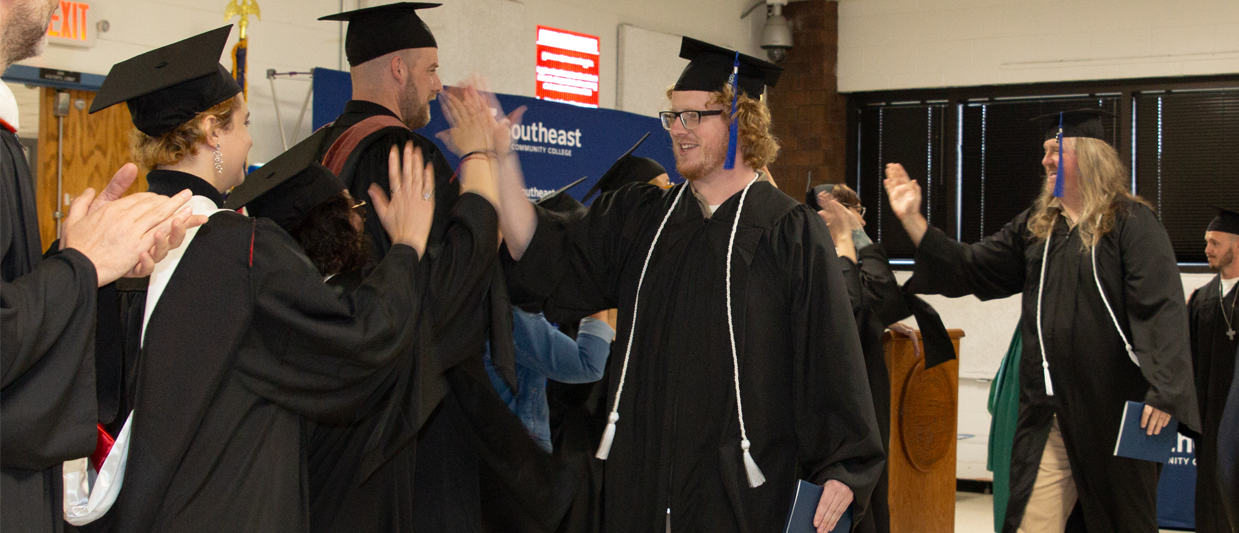 A group of graduates in SCC’s Prison Education Program celebrate with SCC faculty in black caps and gowns, with several people high-fiving and embracing each other. The ceremony was held at the Nebraska State Penitentiary.  