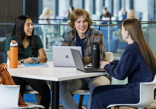From left to right: A woman with long dark hair wearing a dark green top, a male with curly hair wearing a blue and brown plaid shirt, and a woman with long straight hair wearing a navy blue top are sitting around a white round table in a collaborative meeting. The table has a silver MacBook laptop, a black water bottle, and coffee cups. An orange/brown bag is visible on the left side. Large windows in the background show other people working in the bright, contemporary building.