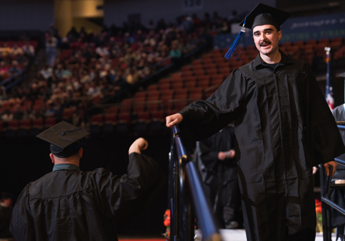 A male graduate in a black cap and gown smiles while walking off the stage at the SCC’s spring graduation, fist-bumping another graduate as they pass.