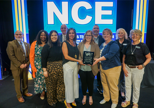 Representatives from Southeast Community College, Central Community College, and Northeast Community College stand in front of a large blue screen displaying "NCE" in big white letters to accept the Outstanding Career and Technical Education Program Award. 