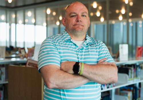 Grant Showalter stands with his arms crossed, wearing a teal and white striped polo shirt and a black smartwatch in the SCC library. He has a serious, confident expression.