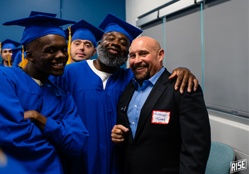 A group of smiling graduates in blue caps and gowns pose with Tommy Moore who is wearing a black blazer and blue button-down shirt wearing a name tag.