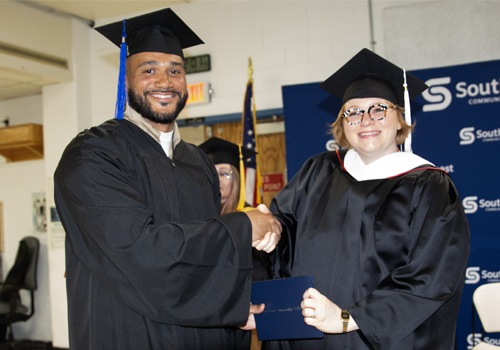 Tammy Zimmer (right) wears a black cap and gown with a white stoll as she hands the diploma to a man in a black cap and gown with a blue tassel at the 2025 prison education graduation ceremony.
