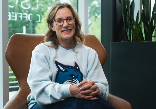 Amy Jurgens is wearing a light gray sweatshirt with a blue bobcat mascot while sitting in a brown chair. Her hands are clasped and she is smiling.