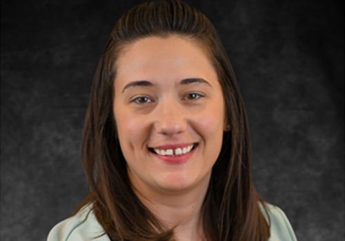 Malisa Kringle has long straight dark brown hair, wearing a light mint-colored top, smiling against a dark gray background.