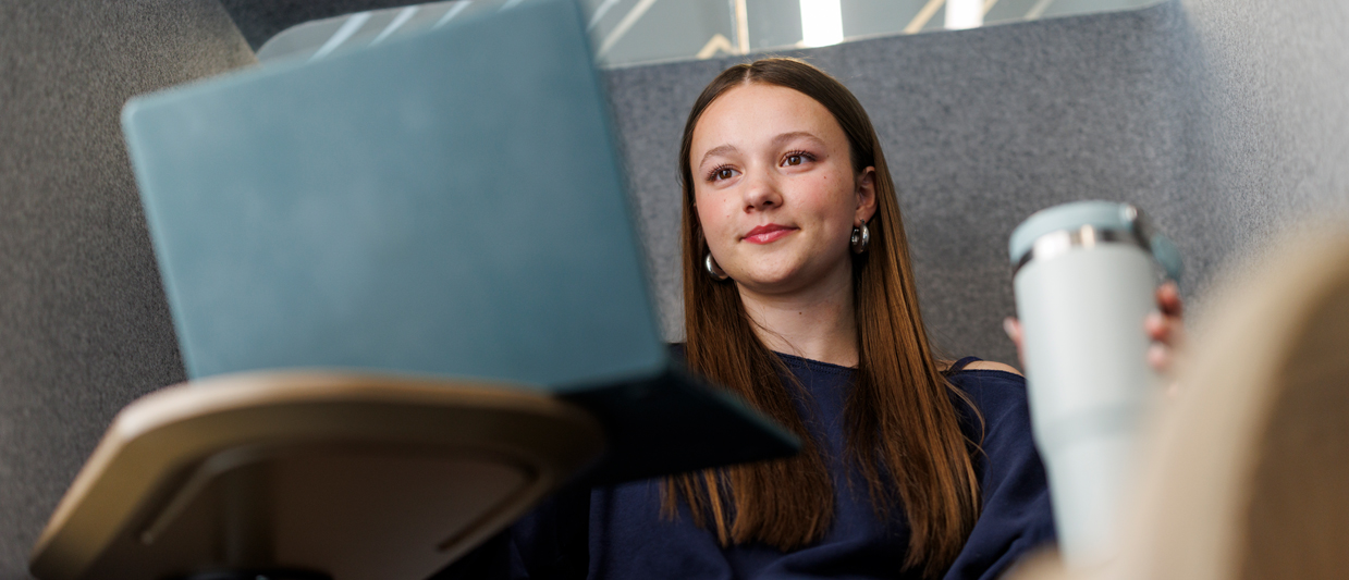 A young woman with long brown hair sits in a light gray chair working on her laptop and holding a cup of coffee, smiling at the camera while wearing a navy blue top.