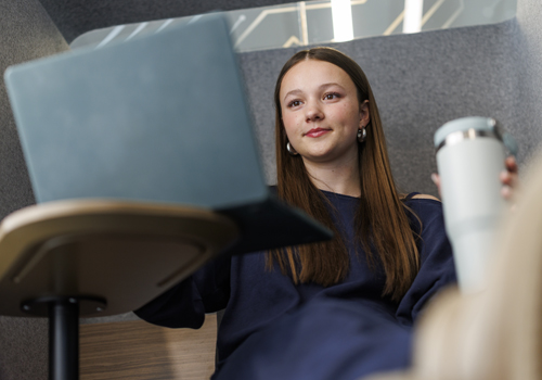 A young woman with long brown hair sits in a light gray chair working on her laptop and holding a cup of coffee, smiling at the camera while wearing a navy blue top.