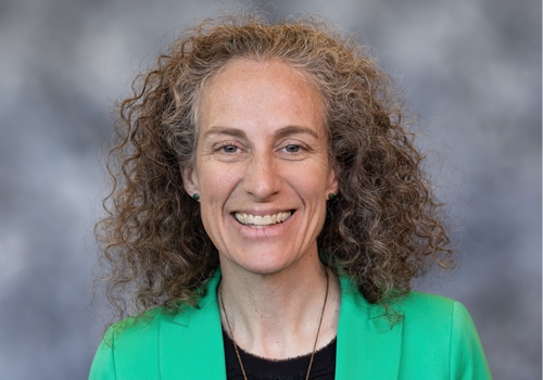 Brooke Hay smiles at the camera wearing a green blazer and black top. 