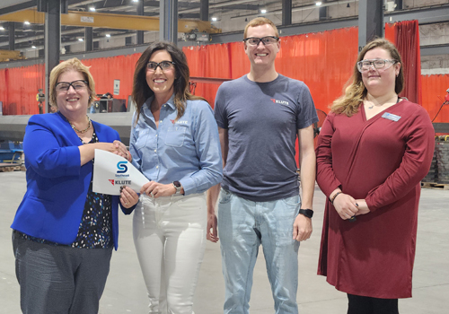 Four professionals standing together in an industrial facility with red safety curtains and manufacturing equipment visible in the background. From left to right: a woman in a blue blazer holding a certificate with an 'S' logo, a woman in a denim shirt, a man in a gray t-shirt, and a woman in a burgundy dress. All are smiling at the camera in what appears to be an award or recognition ceremony.