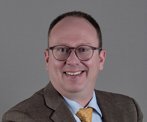 Professional headshot of Matthew Haden wearing round glasses, a brown suit jacket, light blue dress shirt, and yellow patterned tie, smiling at the camera against a gray background.