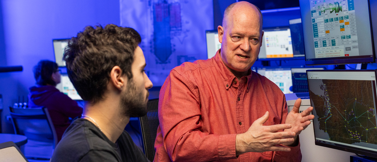 Two men in a blue-lit technical control room having a discussion. The man in a red button-up shirt gestures toward computer monitors displaying technical data and maps while speaking to a colleague in a black shirt. 
