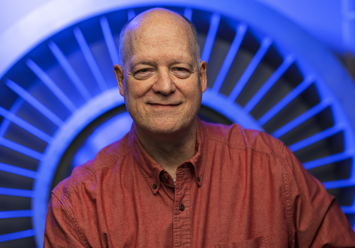 David Madcharo is in front of a blue background, smiling in a brick colored shirt.
