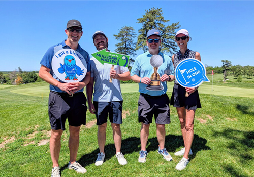 Members of the UBT team pose at Pioneers Golf Course, holding up novelty signs celebrating their achievements: "I Got a Birdie," "Mulligan Master," a trophy, and "Hole in One." They're standing on a green with trees and a fairway in the background on a sunny day.